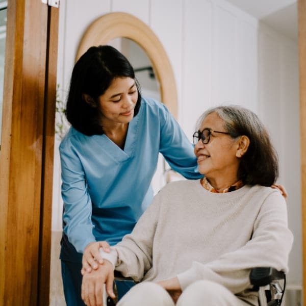 A caregiver in scrubs supporting an elderly woman in a wheelchair at home