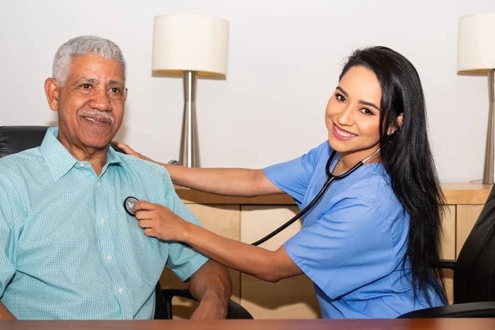 Caregiver in scrubs listening with a stethoscope to a seated senior at home
