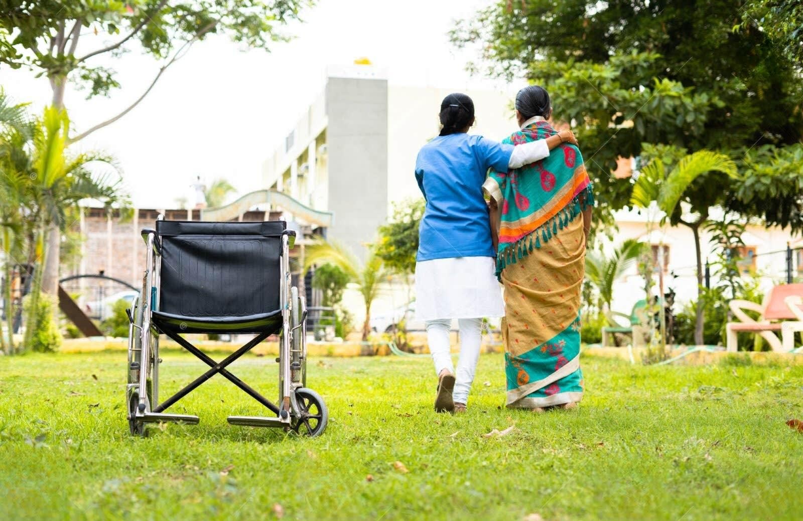 Caregiver supporting a senior walking on a lawn beside a wheelchair, trees and building in the background